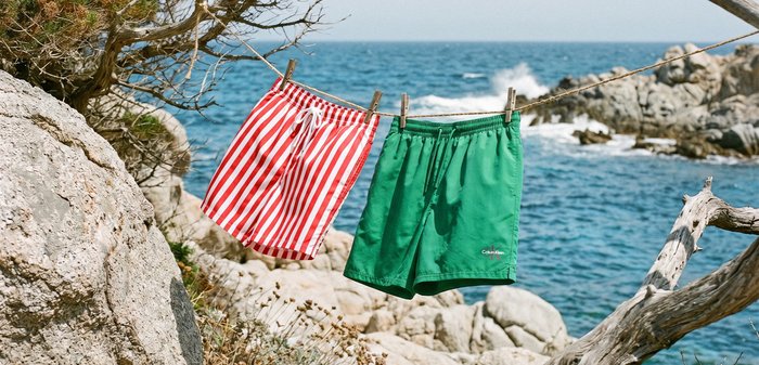 Κόκκινα και λευκά ριγέ και μονόχρωμα πράσινα μαγιό που κρέμονται σε σκοινί για άπλωμα ρούχων δίπλα σε βραχώδη παραλία με κύματα ωκεανού στο φόντο.