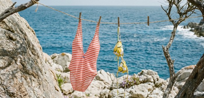 Maillot de bain une pièce rayé et haut de bikini à motif citron suspendus à une corde entre deux arbres au bord d’une côte rocheuse avec la mer en arrière-plan.