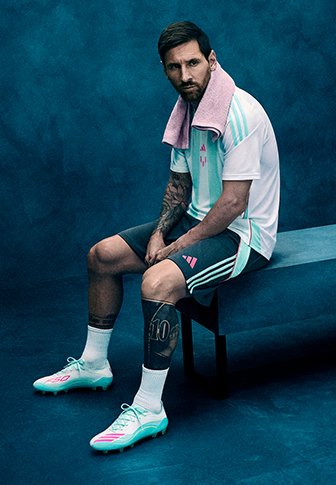 Joueur de football masculin aux bras et jambes tatoués, assis sur un banc, portant un uniforme bleu clair et blanc, des crampons, et une serviette autour du cou.
