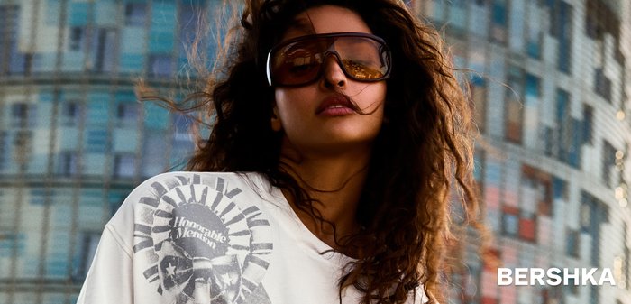 Femeie cu păr creț poartă ochelari de soare supradimensionați și un tricou alb cu nod și design grafic, stând în fața unui fundal cu clădire modernă din sticlă.