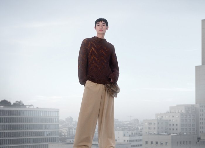Pull en tricot marron avec motif en chevrons. Assorti à un pantalon large beige avec un détail en fourrure sur le côté. Décor urbain.