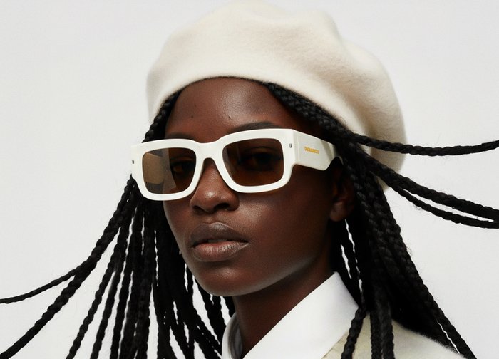 Fehér téglalap alakú napszemüveg barna árnyalatú lencsékkel, krémszínű beretttel és fehér galléros inggel viselve, kiemelve a merész geometriai formát.