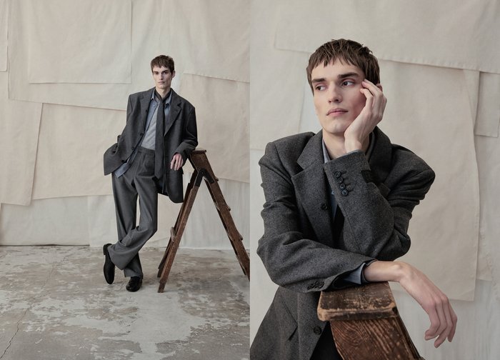 Jeune homme en costume gris posant avec un escabeau en bois sur un fond beige texturé dans un espace de style industriel.