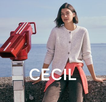 Vrouw in een lichtgrijze cardigan en koraalkleurige trui geknoopt rond de taille leunt op een stenen muur bij rode muntautomatische verrekijkers nabij de zee.