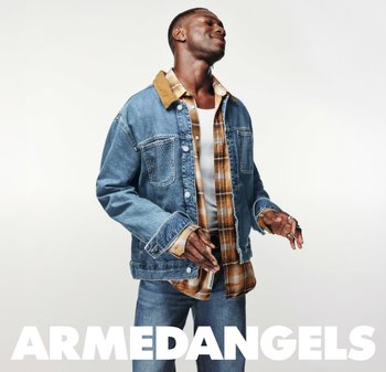 Mann lächelt mit geschlossenen Augen, trägt eine geschichtete Jeansjacke, ein kariertes Hemd, ein weißes Tanktop und Jeans, und steht vor einem weißen Hintergrund.