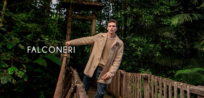 Mand i beige frakke og beige sweater, lænende sig op ad gelænderet på en træbro omgivet af tæt grøn jungleløv under blødt lys.