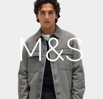 Młody mężczyzna o kręconych ciemnych włosach, ubrany w szarą zapinaną na guziki kurtkę na czarnej koszulce, stojący na tle jasnego, jednolitego tła.