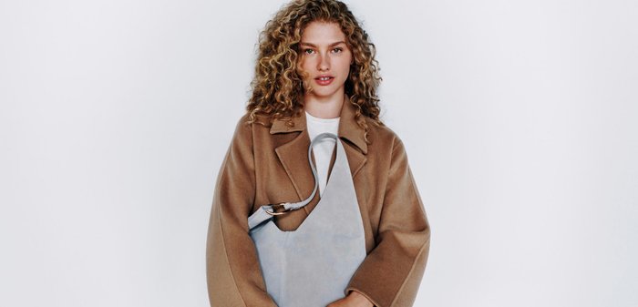 Woman with curly hair wearing a brown coat holds a gray handbag, standing against a white background with Calvin Klein text.