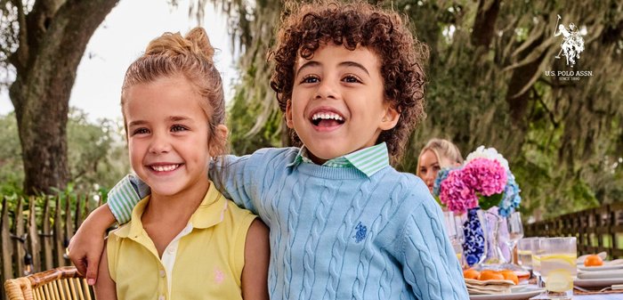 Due bambini sorridenti, una ragazza con una polo gialla e un ragazzo con un maglione blu, con un'area pranzo all'aperto e del verde sullo sfondo.