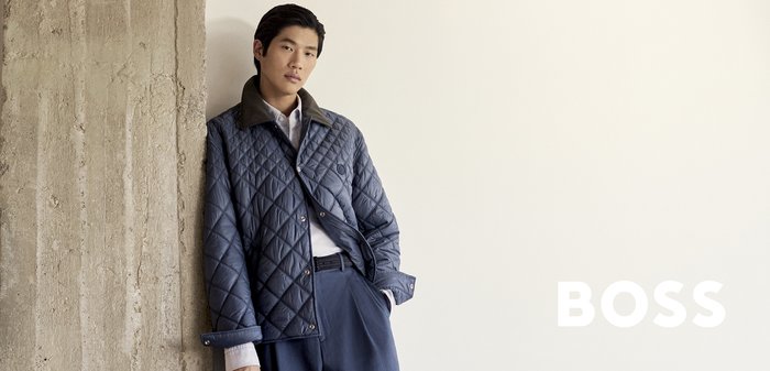 Ein Mann lehnt an einer strukturierten Wand, hält einen braunen Rucksack und trägt eine blaue gesteppte Jacke, ein weißes Hemd und marineblaue Hosen.