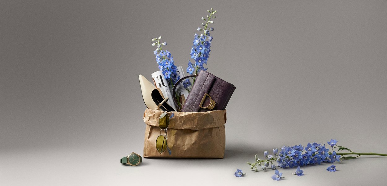 Brown paper bag containing blue flowers, a rolled-up magazine, a white high heel shoe, and a purple clutch, with a green watch and sunglasses nearby.