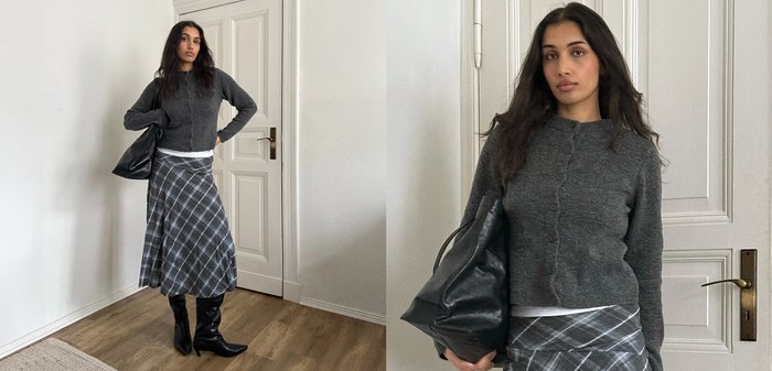 Γυναίκα με γκρι πουλόβερ, καρό φούστα και μαύρες μπότες κρατά μια μεγάλη μαύρη τσάντα, στέκεται σε ένα δωμάτιο με λευκούς τοίχους και ξύλινο πάτωμα.