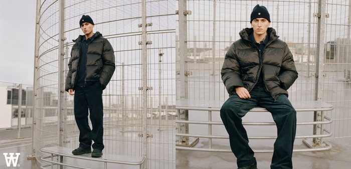 Czarna kurtka puchowa z kapturem, noszona na czarnym topie. Ciemne dżinsy i czarne trampki. Siedzi na metalowej ławce na ogrodzonym terenie.