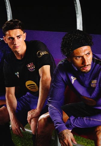 Zwei männliche Fußballspieler sitzen auf einer Bank, tragen FC Barcelona Trainingskleidung, fokussiert und bereit für den Einsatz auf dem Spielfeld.