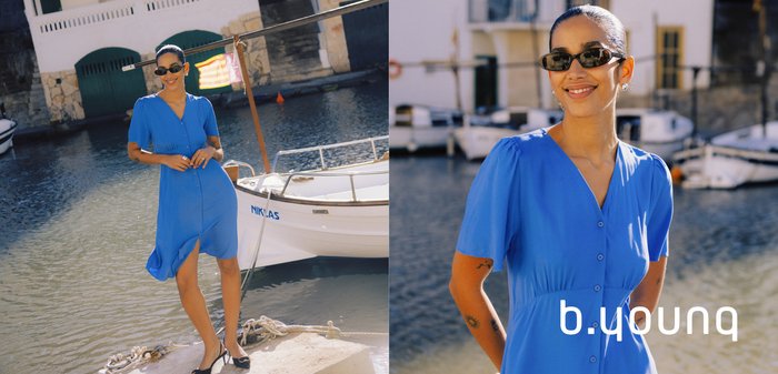 Frau in blauem, geknöpftem Kleid und Sonnenbrille lächelt an einem angelegten Boot an einem sonnigen Tag in der Nähe von Wasser und steinernen Gebäuden.