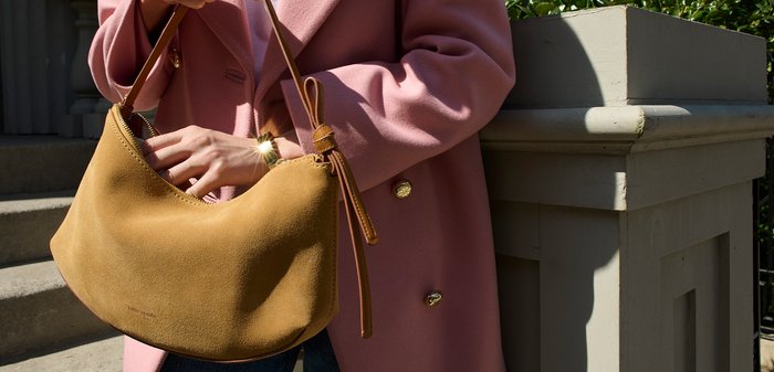 Borsa a tracolla in suede beige con forma curva e una singola tracolla. Si nota la superficie testurizzata e un design minimalista, abbinata a un cappotto rosa.