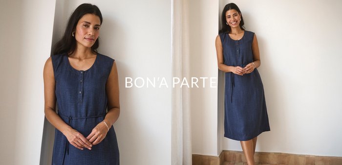 Žena sa usmieva a opiera sa o bielu stenu, oblečená v tmavomodrých šatách bez rukávov, s dĺžkou po kolená, a čiernych balerínach.