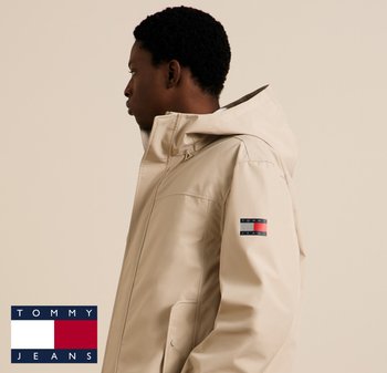 Beżowa wodoodporna kurtka z kapturem, wykonana z gładkiego materiału. Na lewej rękawie znajduje się naszywka z logo w kolorach czerwonym, białym i niebieskim.