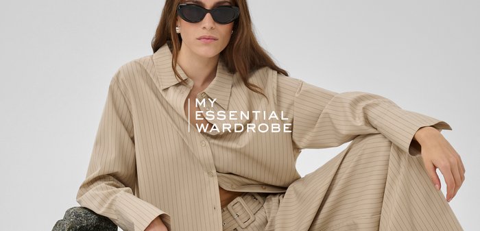 Naine, kes kannab beežika triibulise mustriga üle suuruses bleiserit ja laia lõikega pükse, musta kassi-silmadega päikeseprille, toetudes tumeda tekstuurse kivi vastu.