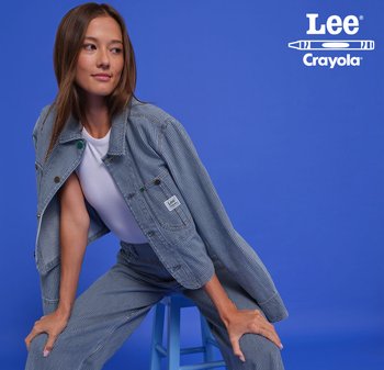 Veste en denim rayée bleue avec des accents verts et un patch de logo, portée sur une chemise blanche. Le modèle pose assis sur un tabouret bleu clair.