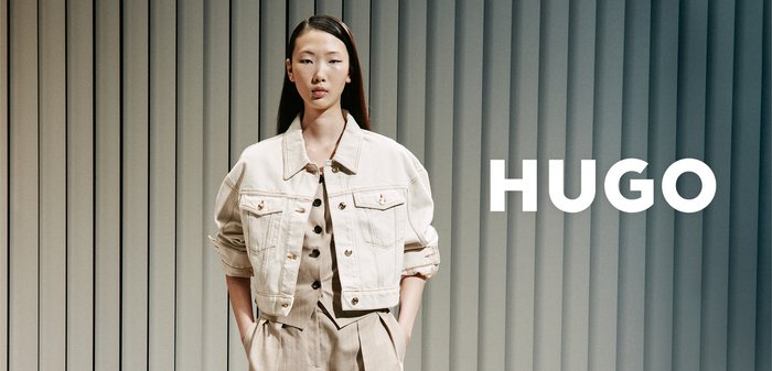 Modelo femenina con chaqueta beige y pantalones a juego delante de persianas verticales con texto "HUGO" a la derecha.