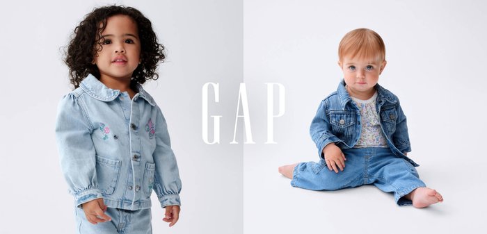 To småbørn iført blå denimjakker og jeans, den ene stående og den anden siddende, mod en hvid baggrund med teksten "GAP" i midten.
