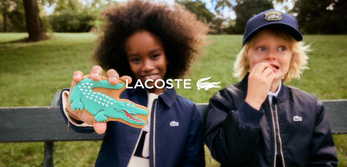 Two children wearing navy jackets sit outdoors; one holds a green alligator-shaped cookie, the other eats a snack.