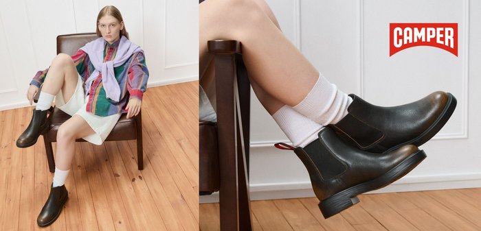 Bottines Chelsea en cuir noir avec un léger brillant, présentant un empiècement en cuir marron contrastant à l'avant et une languette rouge. Portées avec des chaussettes blanches.