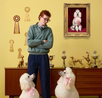 Ung man som står med två vita pudlar som bär rosa rosetter, omgiven av hundtroféer och band på en gul vägg och skåp.