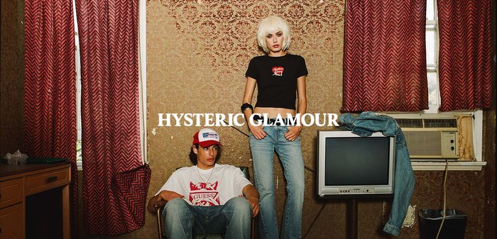 Młody mężczyzna w swobodnych ubraniach i czapce siedzi obok stojącej kobiety z blond bobem, oboje w dżinsach, na tle vintage tapety z telewizorem obok.