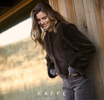 Bruine, fuzzy cropped jas met lange mouwen, van textiel met structuur. Draagt over een blauw-grijs shirt, gecombineerd met grijze jeans. Natuurlijke houten achtergrond.