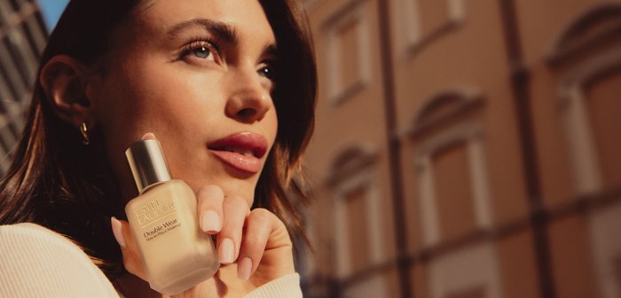 Mujer sosteniendo una botella de base Estée Lauder Double Wear cerca de su cara, con luz cálida del sol y la fachada de un edificio en el fondo.
