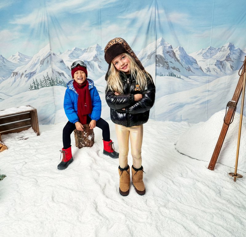 Divi bērni ziemas apģērbā, viens zilā mētelī, sarkanā šallē un sarkanos zābakos, otrs melnā pūkainā jaku un bēšos zābakos, stāv uz sniega.