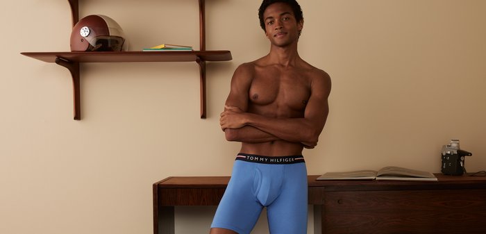 Blaue stretch Baumwoll-Boxershorts mit einem schwarzen Bund, der rote und weiße Streifen hat. Das Modell steht vor einer hellbeigen Wand.
