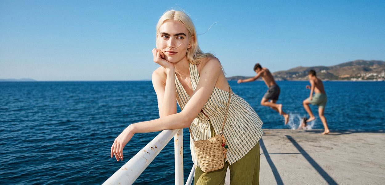 Jeune femme aux longs cheveux blonds appuyée sur une rambarde au bord de la mer, deux garçons sautant dans l'eau depuis un ponton en arrière-plan.