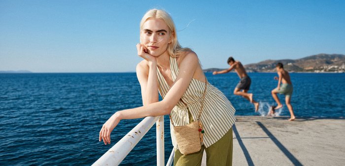 Jeune femme aux longs cheveux blonds appuyée sur une rambarde au bord de la mer, deux garçons sautant dans l'eau depuis un quai en arrière-plan.