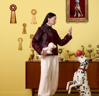 Donna che tiene un cane di peluche e fa un segno con la mano mentre un dalmata fa equilibrio con una palla rossa sul naso in una stanza piena di trofei con nastri per cani.