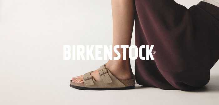 Braune Birkenstock-Sandalen mit zwei verstellbaren Riemen und einer strukturierten Oberfläche, neben einem Bein, das einen dunklen Rock trägt, auf einem hellen Hintergrund.