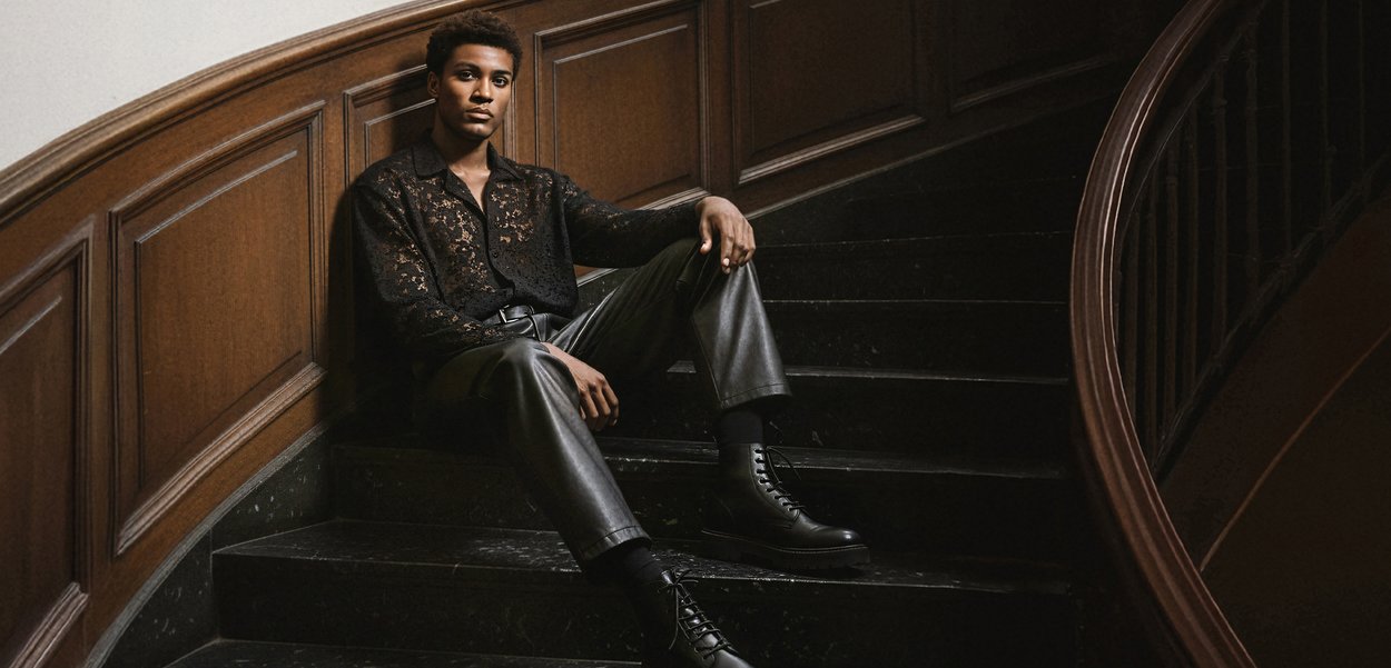 Model așezat pe o scară curbată, purtând o cămașă din dantelă neagră, pantaloni din piele și cizme negre cu șireturi. Panouri de lemn și trepte din piatră întunecată.