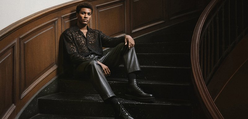 Mannequin assis sur un escalier courbé, portant une chemise en dentelle noire, un pantalon en cuir et des bottes noires à lacets. Paroi en bois et marches en pierre sombre.