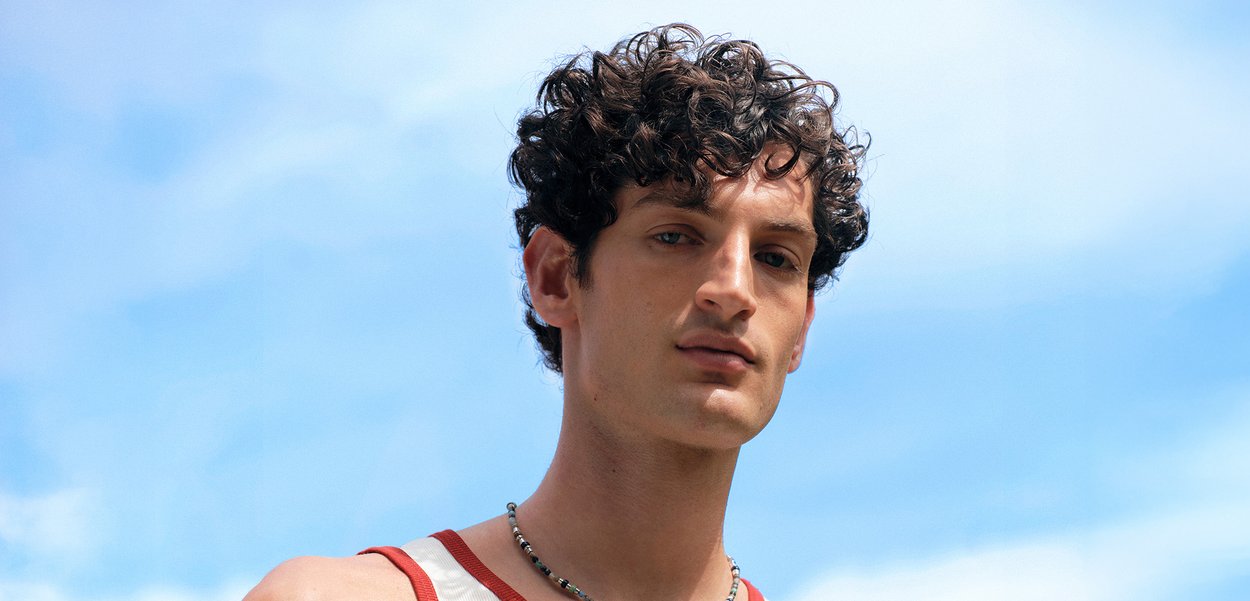 Giovane uomo con capelli ricci e scuri che indossa una canotta senza maniche e una collana, guarda con sicurezza la fotocamera contro un cielo azzurro limpido.