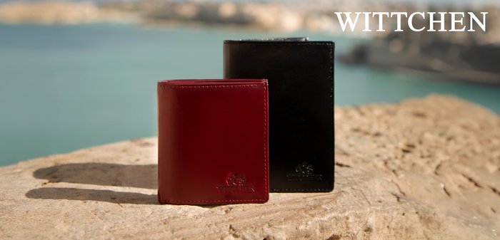 Zwei Lederbrieftaschen, eine rote und eine schwarze, stehen aufrecht auf einer Steinoberfläche neben einem Gewässer mit unscharfem Hintergrund.