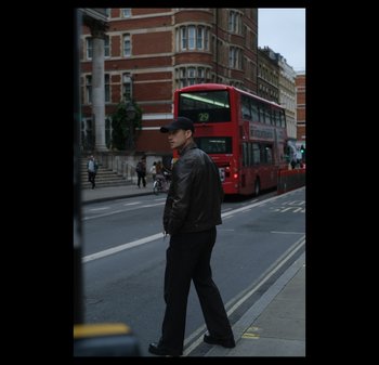 Un uomo con un cappello nero e una giacca di pelle marrone si trova su una strada di città. Dietro di lui si vede un autobus a due piani rosso, con edifici in mattoni nelle vicinanze.