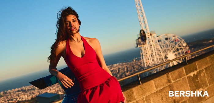 Mlada žena u crvenoj halter haljini naslonjena na plavi teleskop, s pogledom na grad i ocean u pozadini tijekom zalaska sunca.