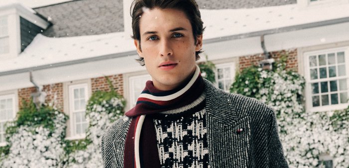 Junger Mann im Freien im Winter, der einen gemusterten Pullover, einen grauen Mantel und einen gestreiften Schal trägt, mit Schnee und einem Haus im Hintergrund.