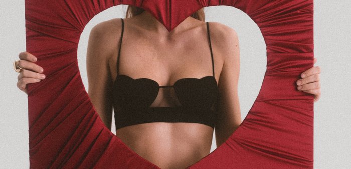 Mujer con sujetador negro sostiene tela roja con un recorte en forma de corazón que revela la parte superior de su cuerpo sobre un fondo claro.