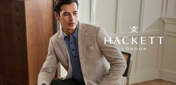 Vīrietis, tērpies bēšā žaketē, zilā krekliņā un tumši zaļos biksēs, sēž krēslā istabā ar koka mēbelēm un paneļotu sienu.