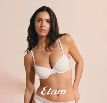 Vrouw met lang donker haar, gekleed in een witte lingerieset, poserend tegen een effen beige achtergrond, merknaam "Etam" zichtbaar.