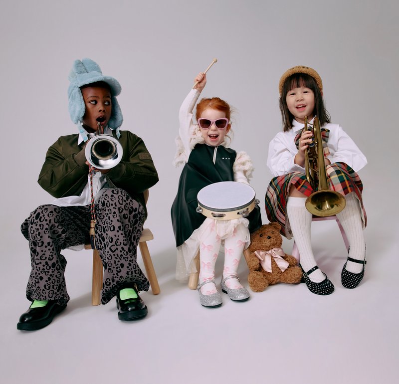 Tre bambini seduti su sgabelli di legno. Uno suona una tromba, un altro tiene un tamburello e il terzo suona una tromba, tutti vestiti con abiti colorati.