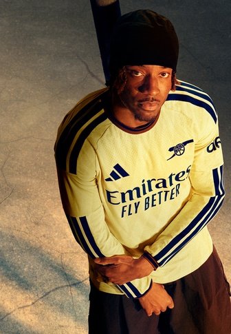 Mann trägt ein gelbes Arsenal-Fußballtrikot mit Adidas- und Emirates-Logos, steht mit verschränkten Armen und schaut in die Kamera.